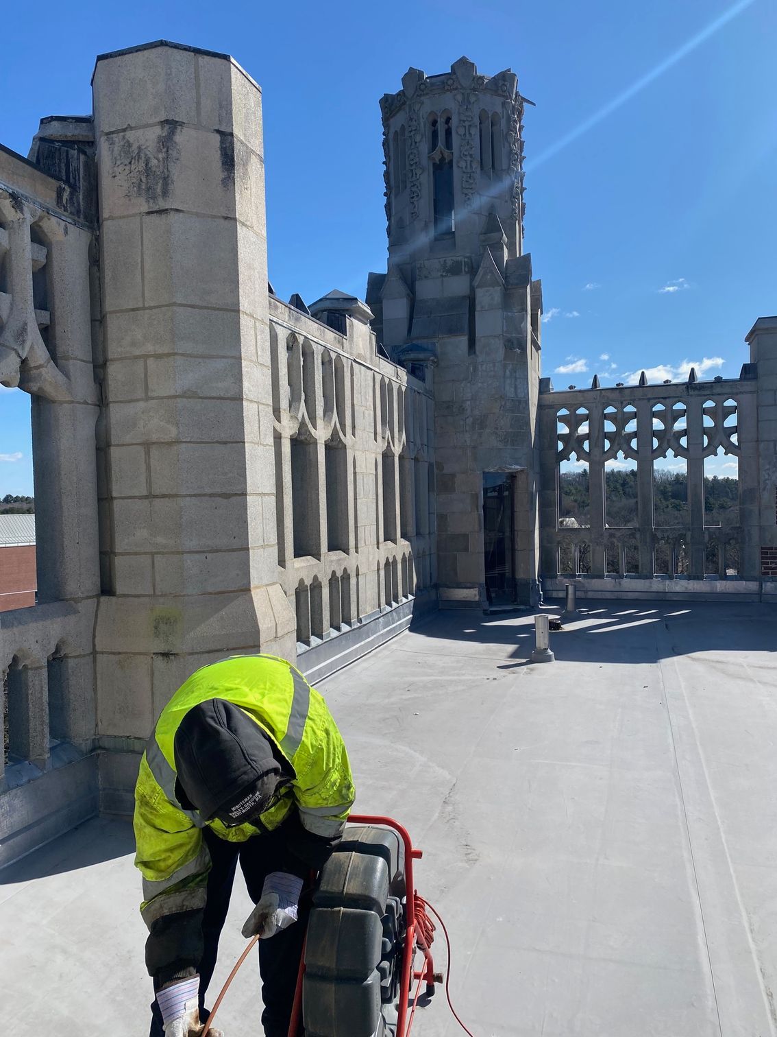 Roofer in neon yellow vest repairs a flat roof with a stone building and blue sky in the background.