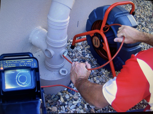 Plumber inspecting sewer pipe with camera. Red shirt, blue equipment, white pipe, outdoors.