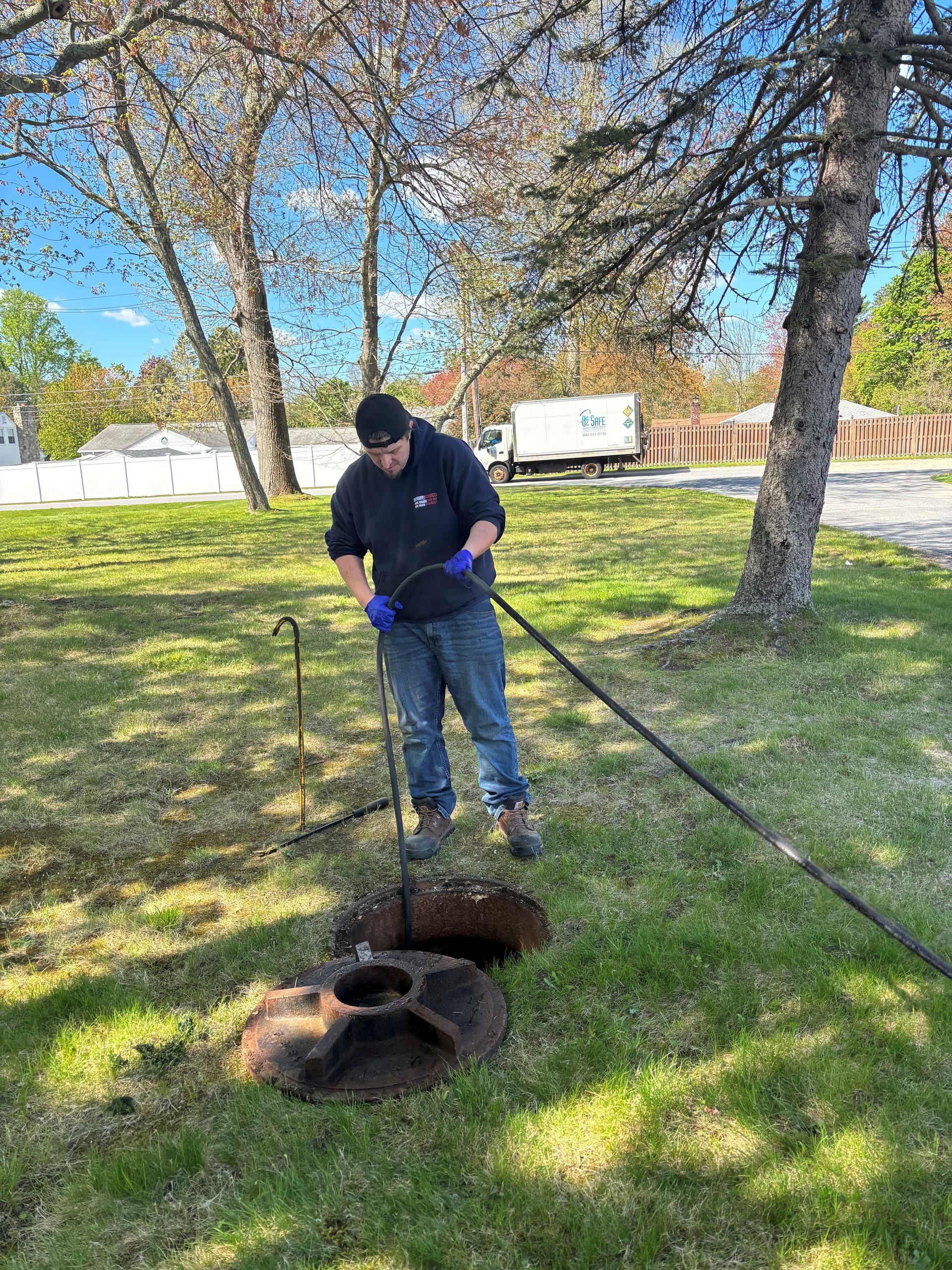 Person using a long tool near an open ground hole in a grassy park with trees nearby.