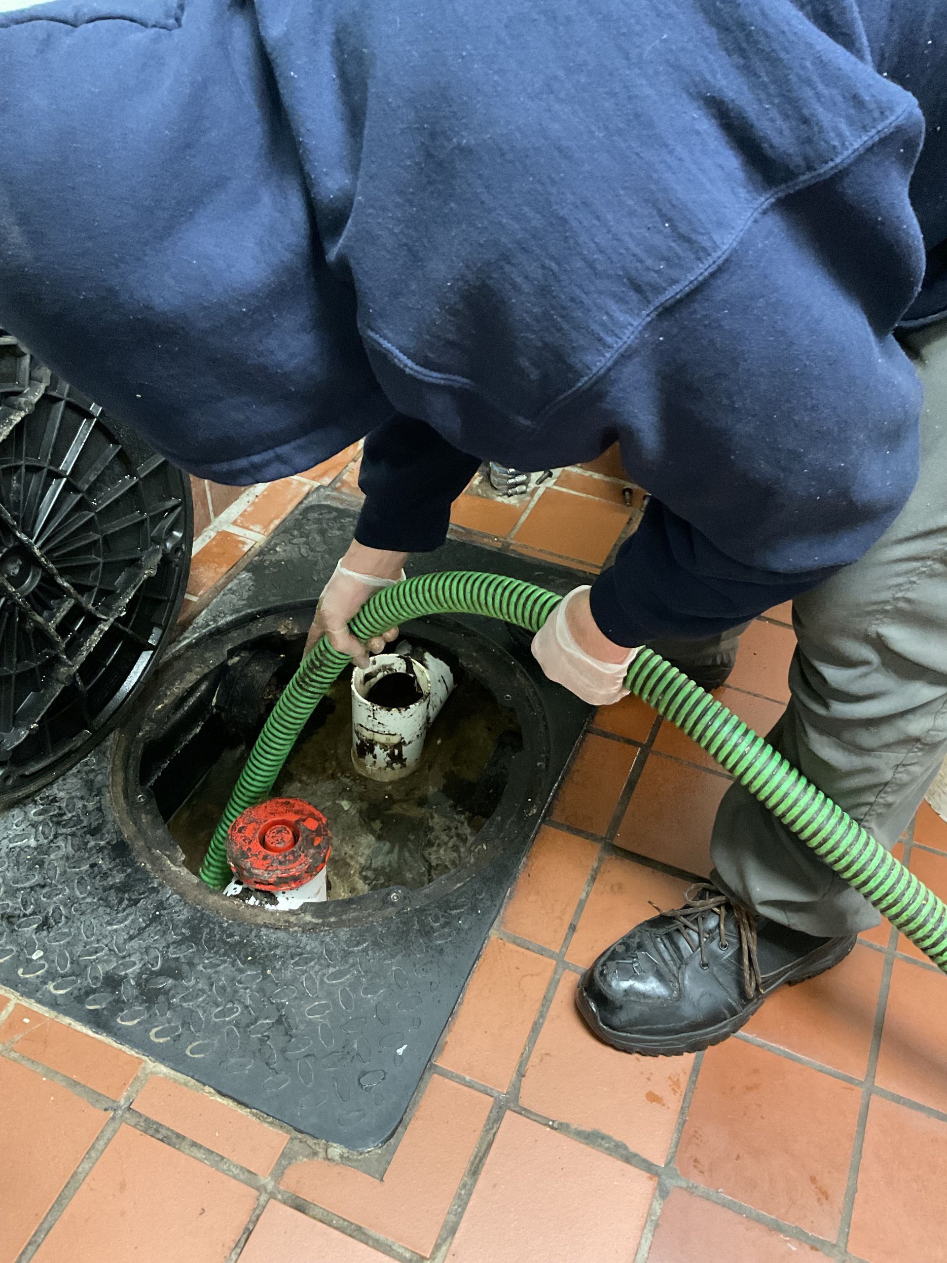 Person in blue sweater, pumping liquid from a grease trap on a tile floor.