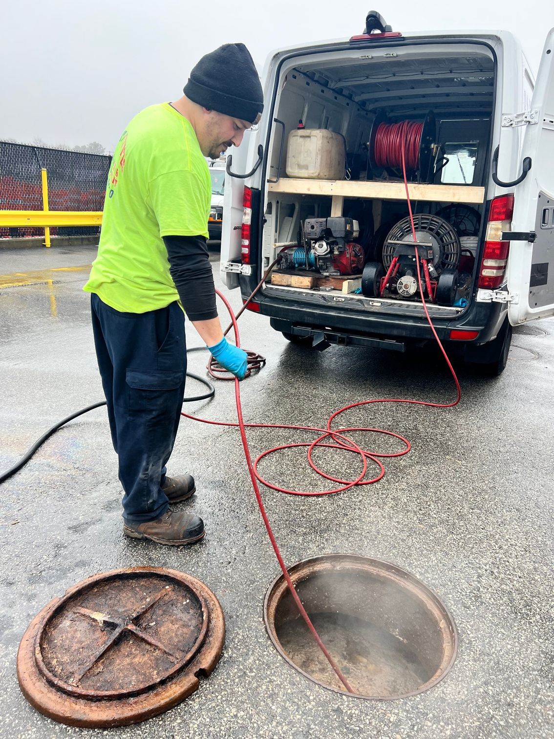 Man in green shirt working on a sewer line with equipment in a van. Manhole cover open on the ground.