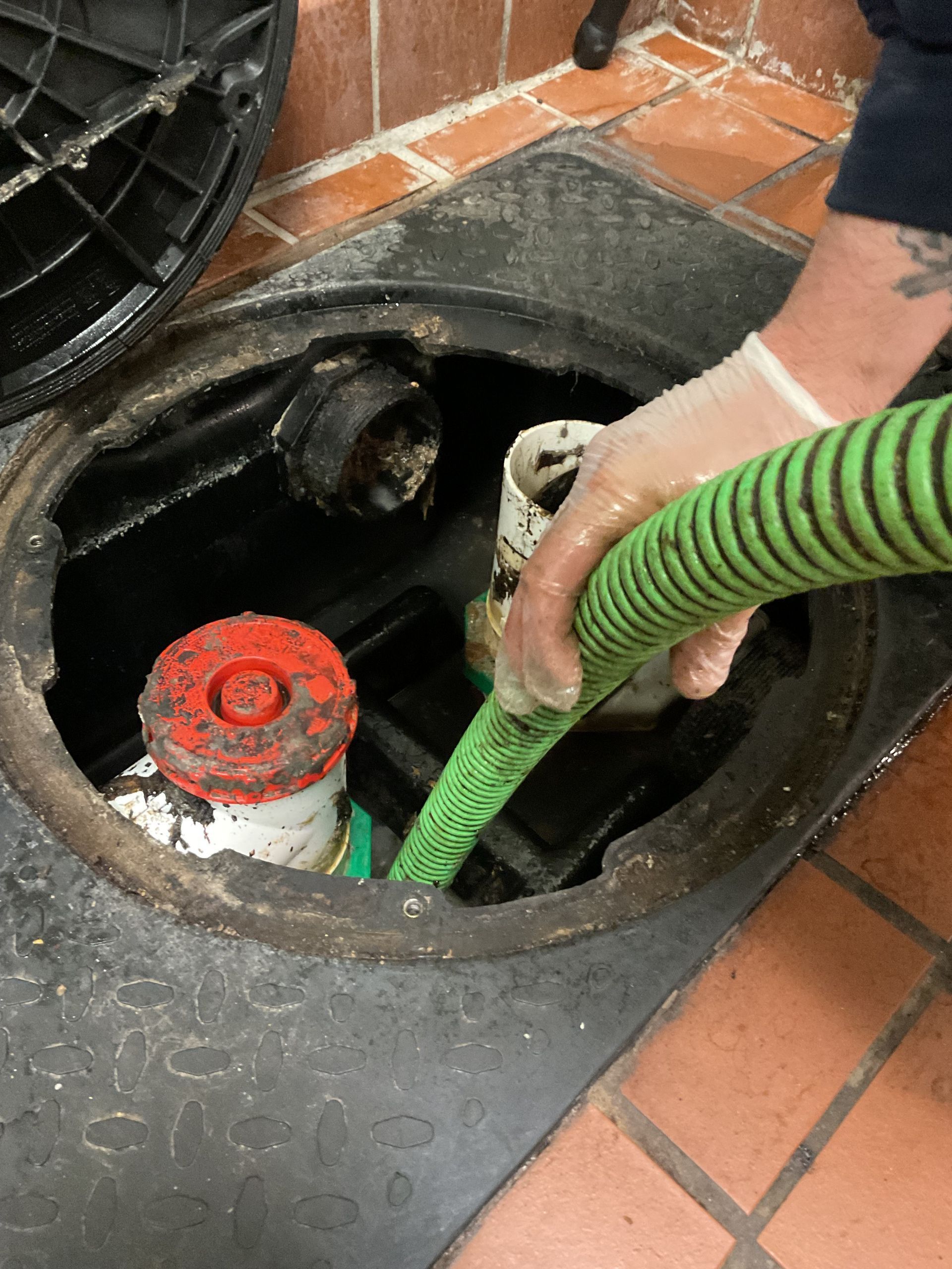 Person in gloves with a green hose, cleaning a grease trap in a commercial setting.