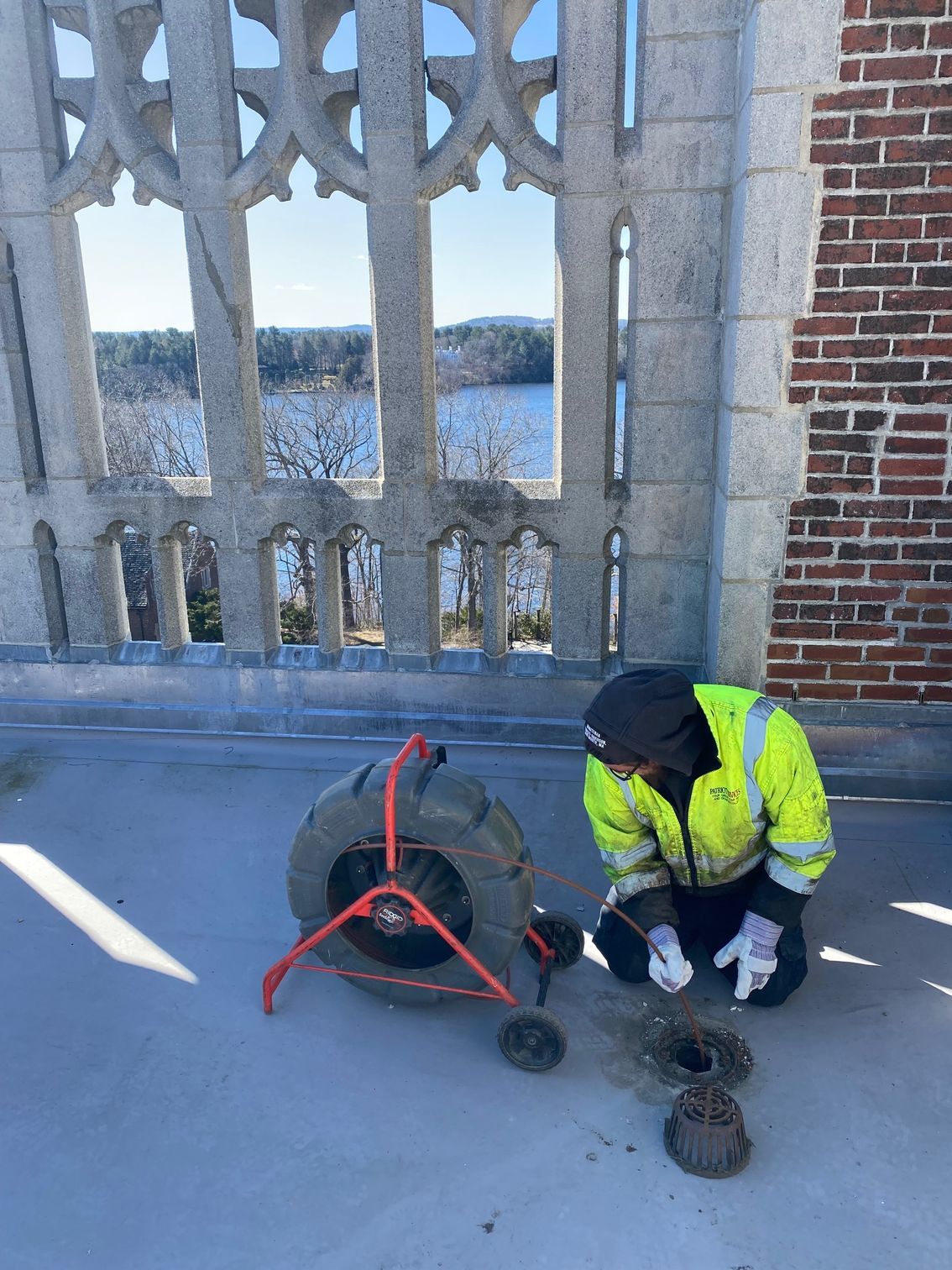Worker in safety vest examines equipment on a rooftop, with water and a decorative window in the background.