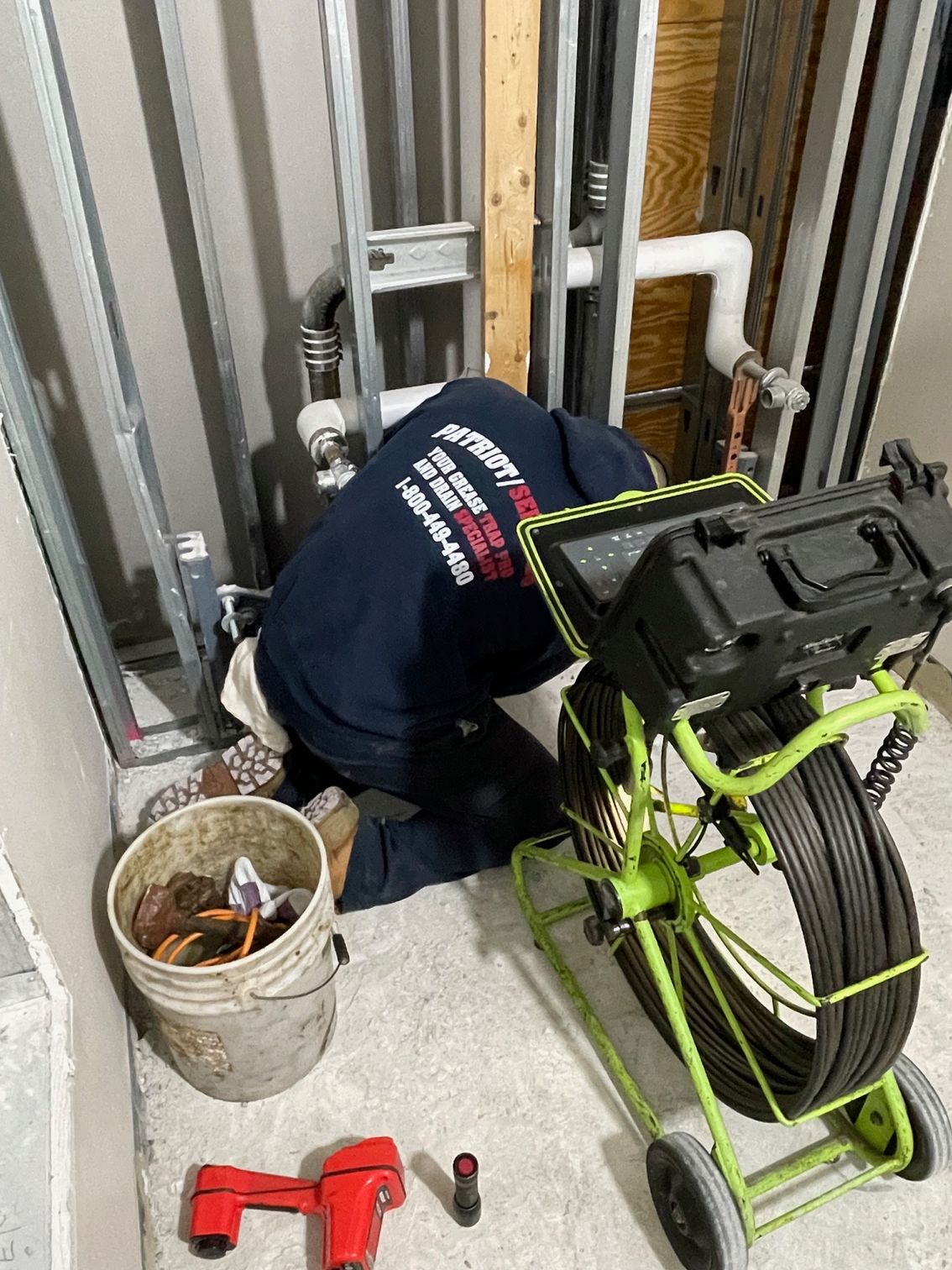 Plumber kneeling, inspecting pipes near a wall with inspection camera on wheels and bucket.