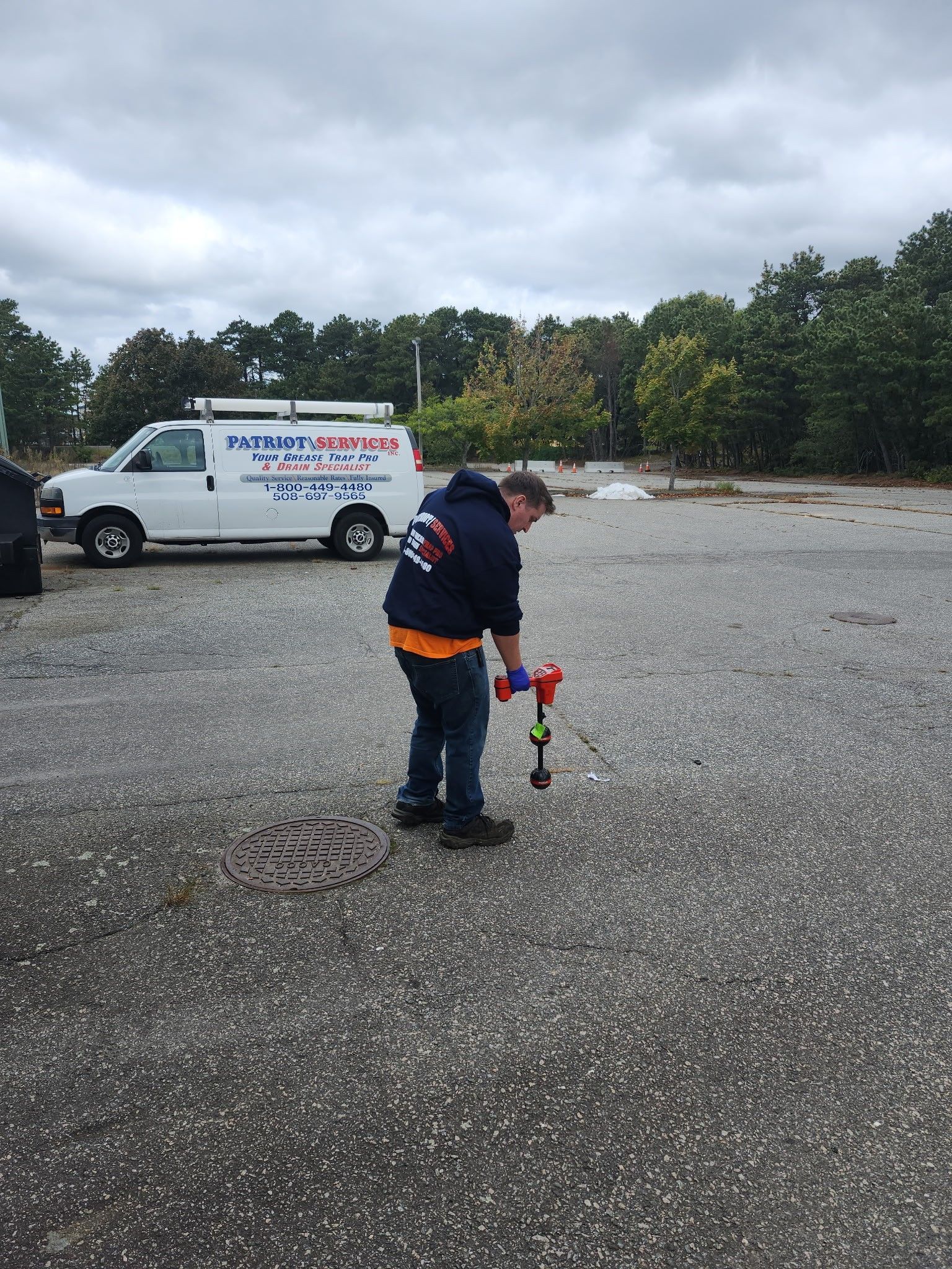 A plumber in blue and orange works on a manhole in a parking lot, with a white van nearby under a cloudy sky.