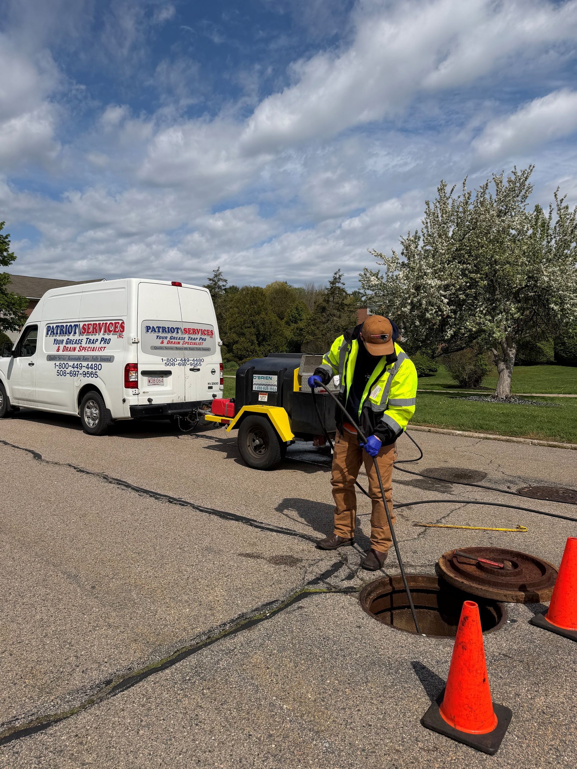 A Patriot Services worker in a high-vis jacket working near cones and cables beside a white van on a paved lot.