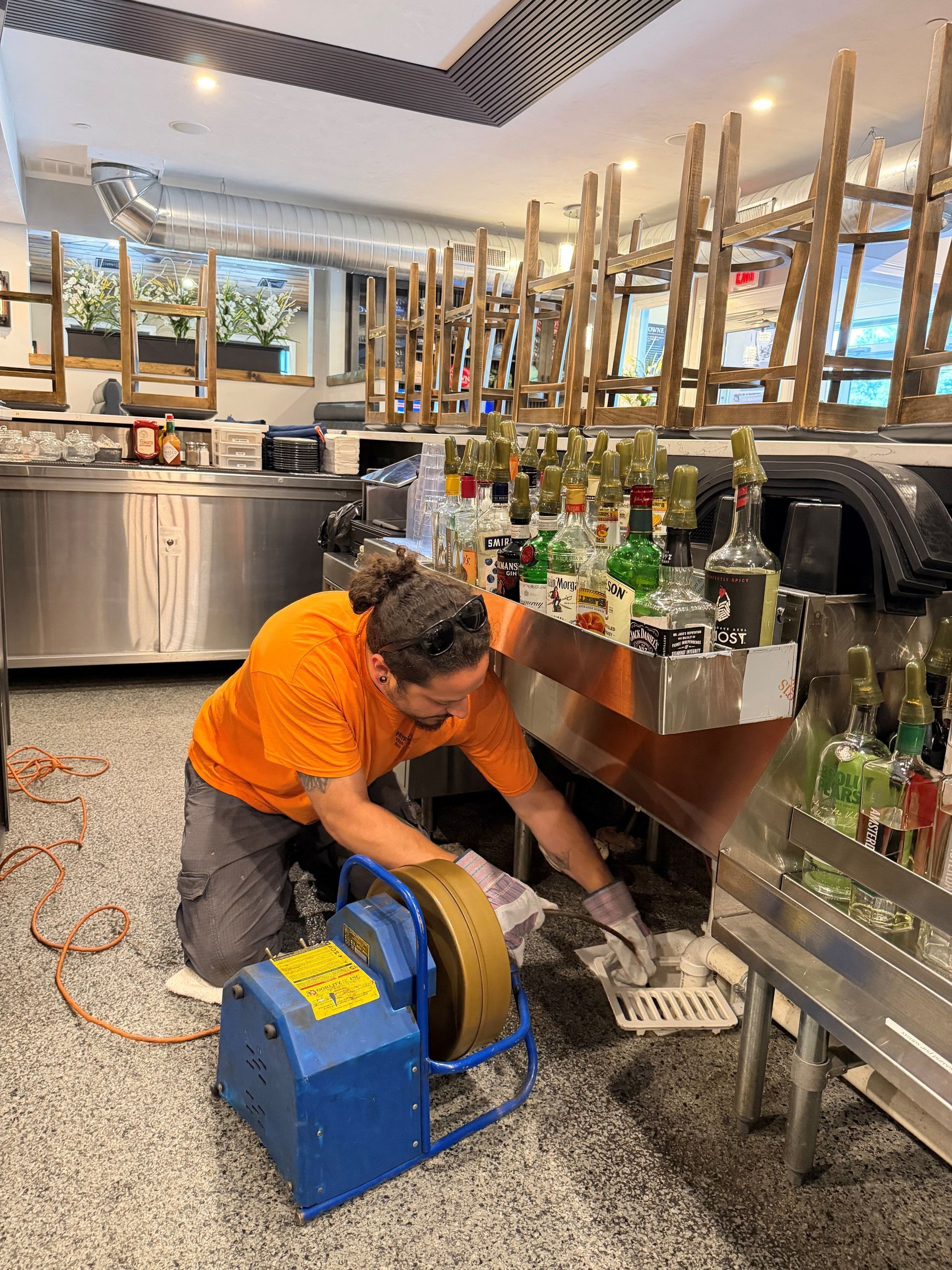 Worker in orange uniform cleaning under a bar counter with a blue floor machine in a modern restaurant.