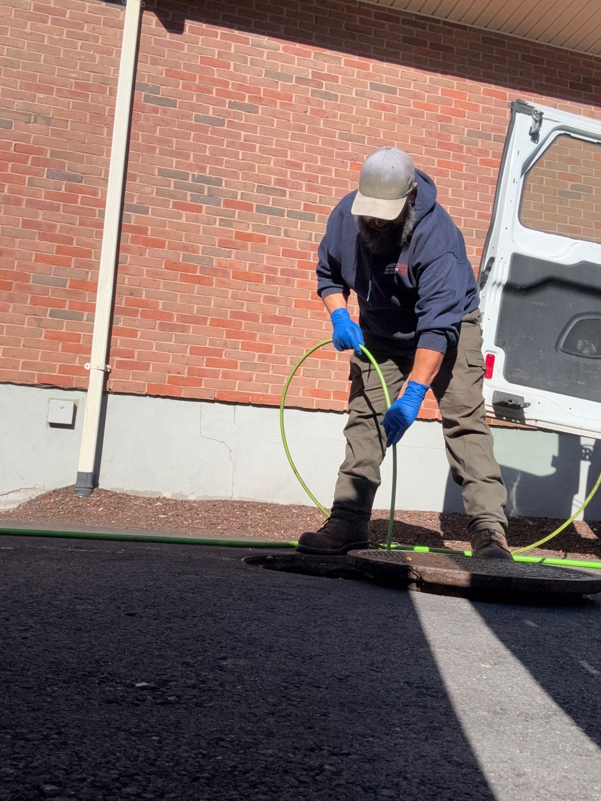 Man in blue gloves and hoodie using a hose on a dark surface near a brick wall and van.